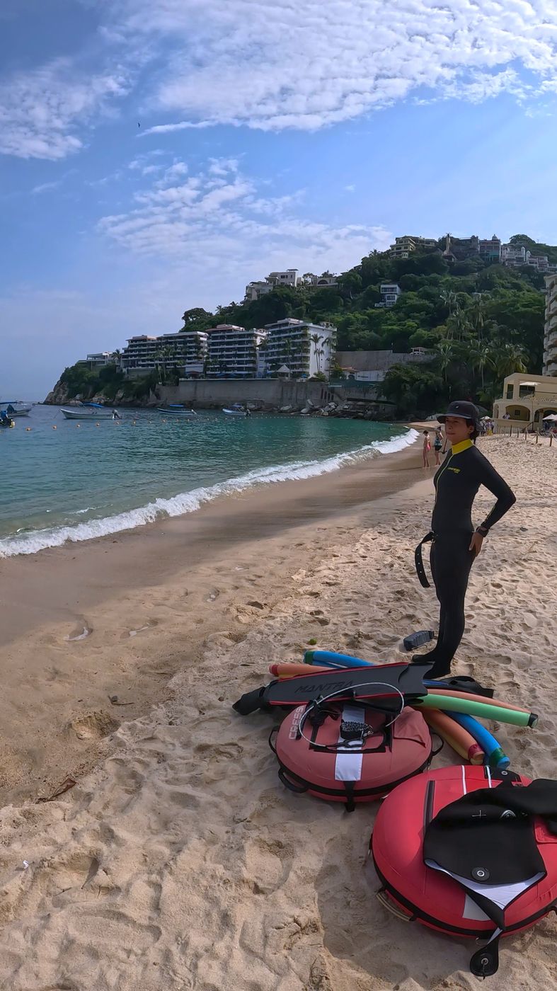 Apneísta con traje de neopreno de pie en la arena junto a boyas rojas y aletas, con hoteles en el cerro al fondo
