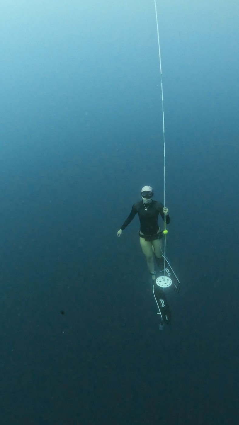 Apneísta con traje oscuro y capucha blanca en el plato de fondo del cabo, en agua verde azulada profunda