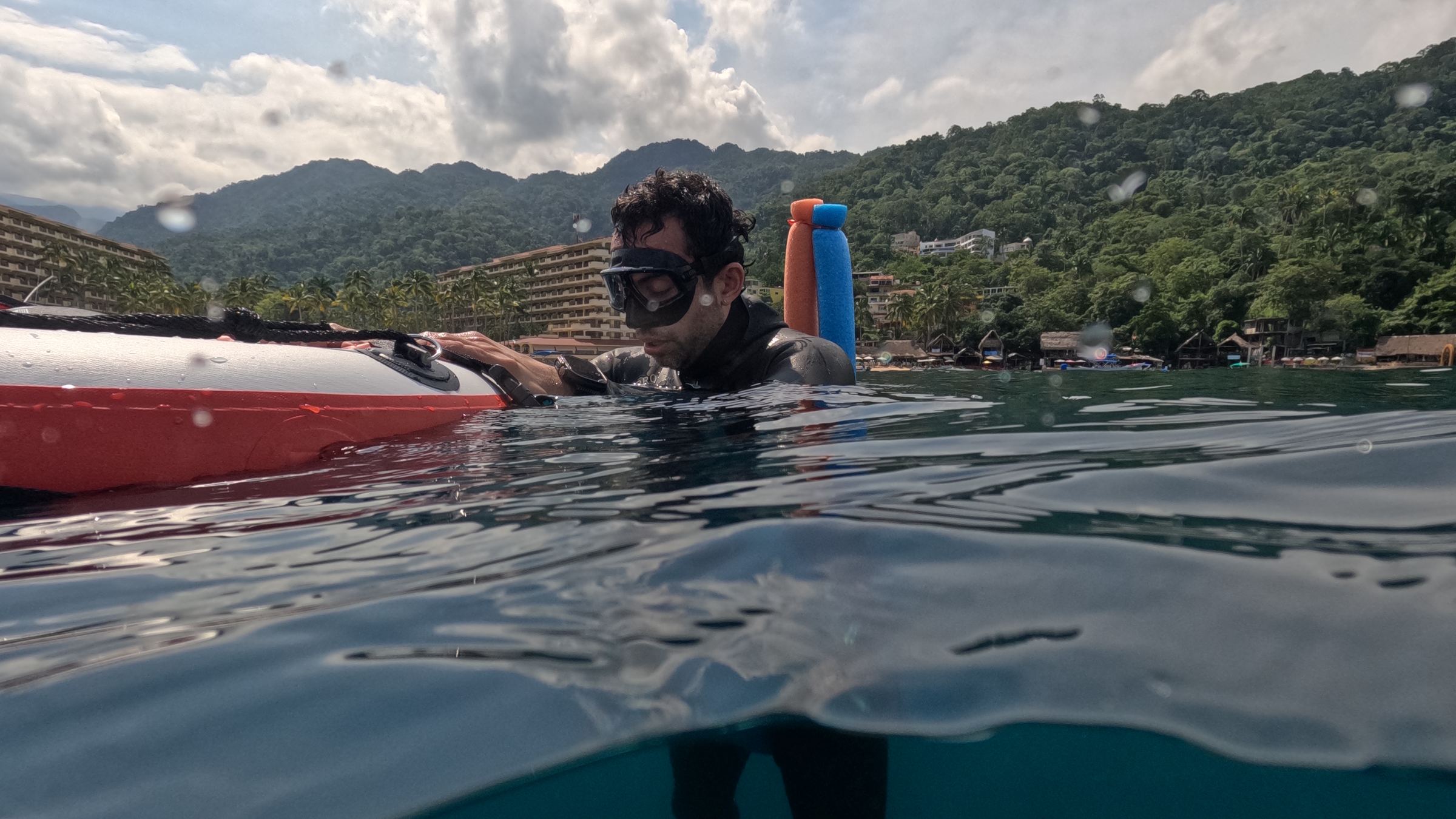 Apneísta con visor descansando sobre una boya roja en la superficie, con hoteles y montañas verdes al fondo