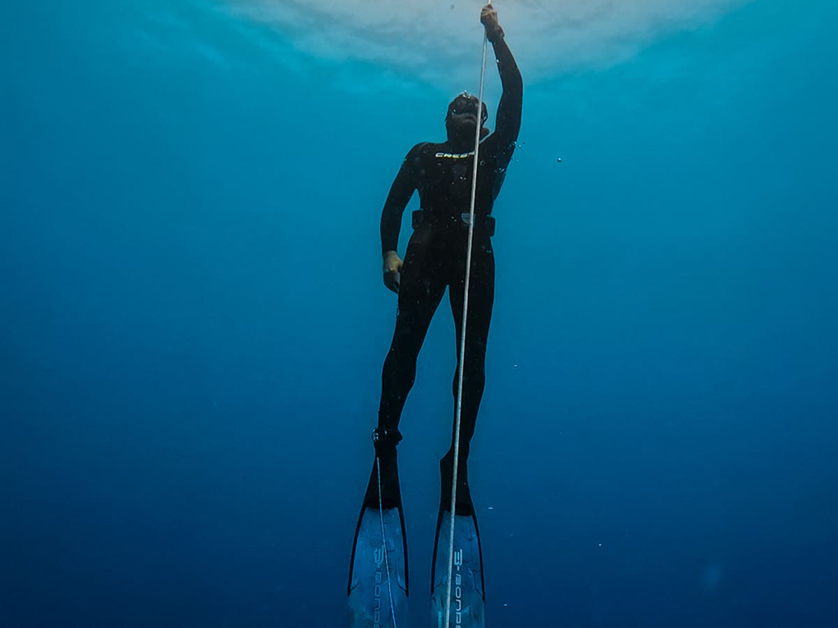 Freediver in a black wetsuit pulling up a line toward the bright surface