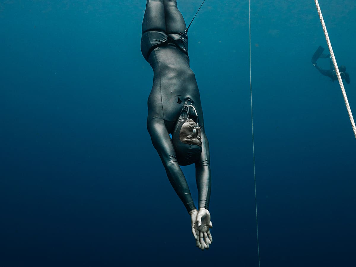 Freediver in a smooth dark suit gliding head-first down beside the line