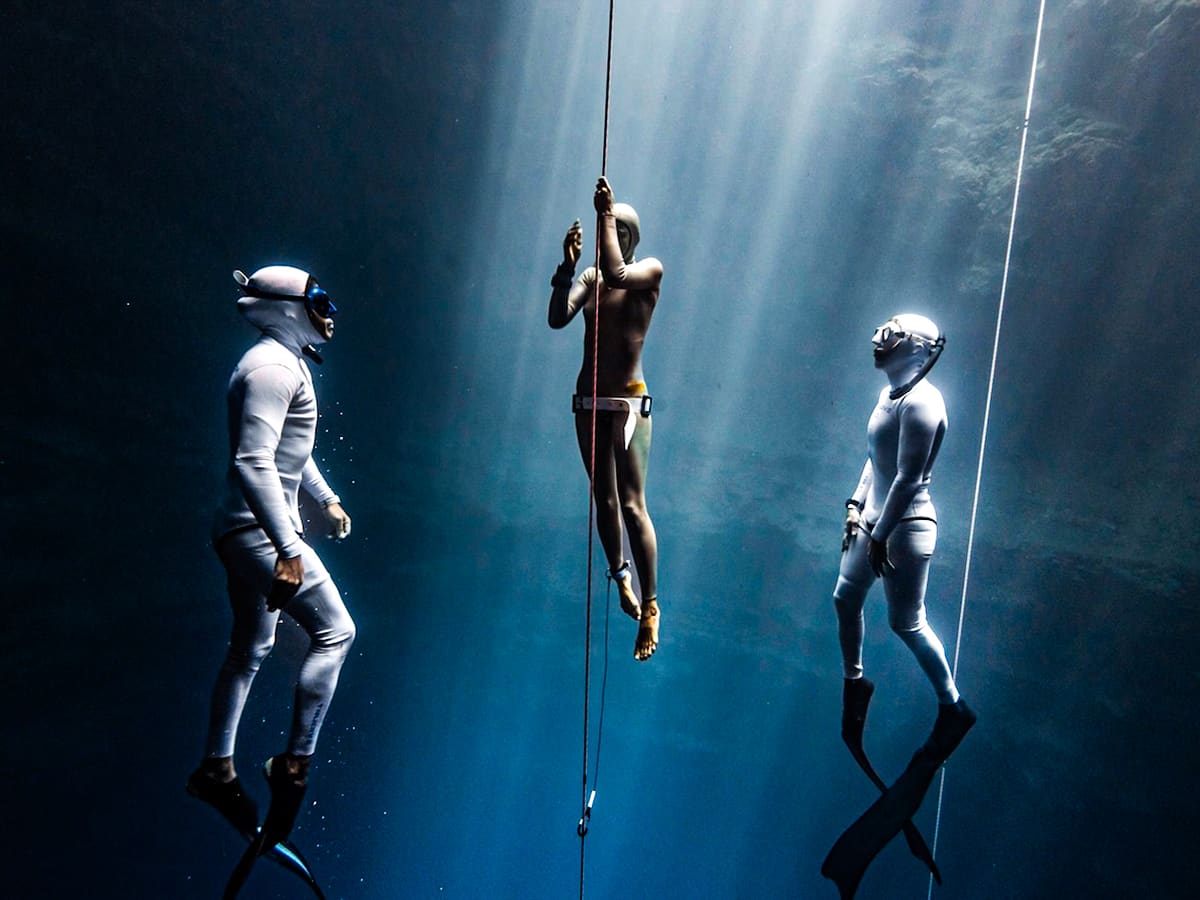 Three freedivers on lines under shafts of light, two in white suits watching the one in the middle