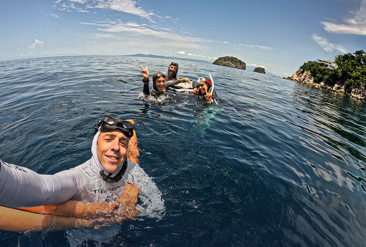 Apneísta con capucha blanca tomándose una selfie en el mar, con otros tres apneístas e islotes al fondo