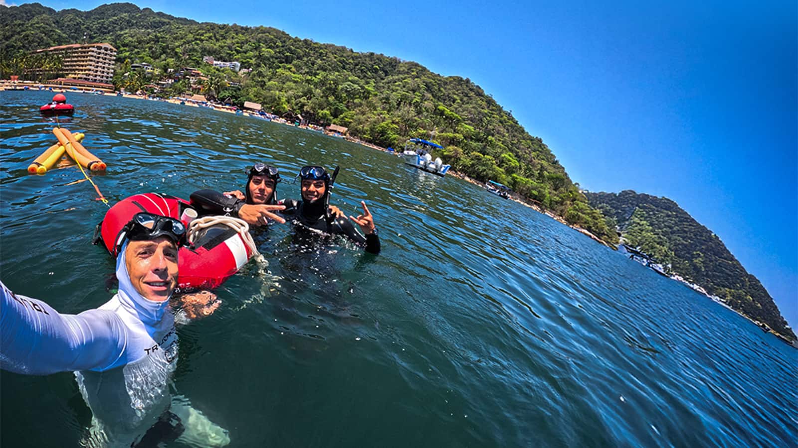Tres apneístas en una boya roja tomándose una selfie, con playa y cerros de selva al fondo