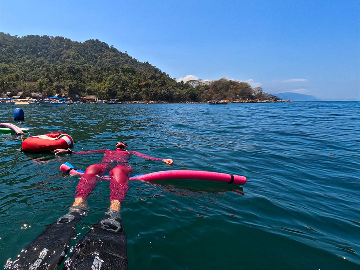 Apneísta con traje rosa flotando sobre un tubo rosa junto a una boya roja, con playa y cerros al fondo