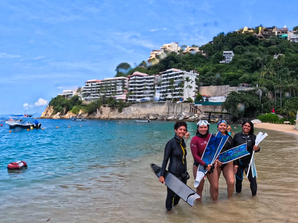 Four freedivers in wetsuits holding long fins in the shallows, hillside hotels behind