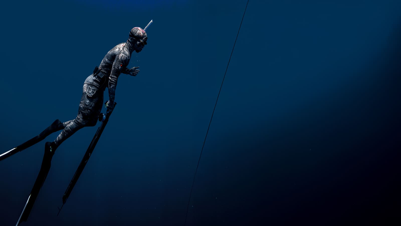Spearfisher in a camouflage wetsuit diving down with a speargun in dark blue water