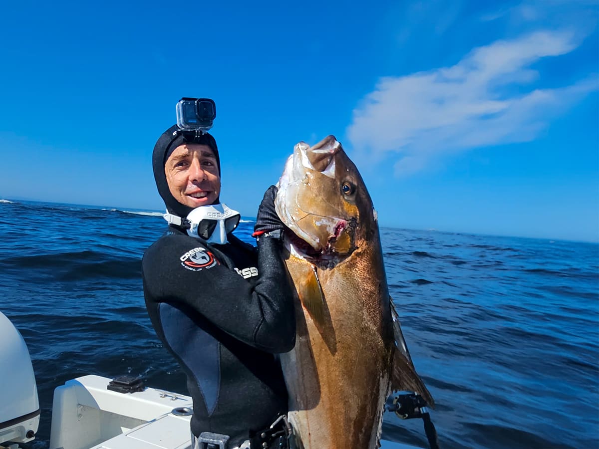 Pescador submarino con capucha y cámara en la cabeza mostrando un pez grande en la lancha